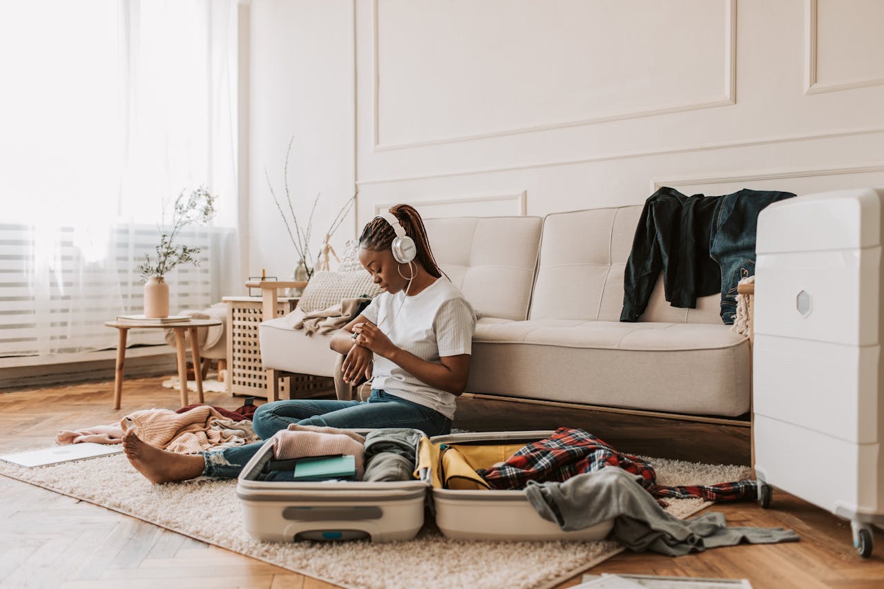 gallery-03 Young woman sits at home packing a suitcase while listening to music, surrounded by clothing items.